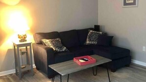 A living room with a dark-colored sofa, two patterned throw pillows, a small side table with a lamp, and a red tray on a wooden coffee table. - Townhomes Near the Bay - #1 (Grand Bay)