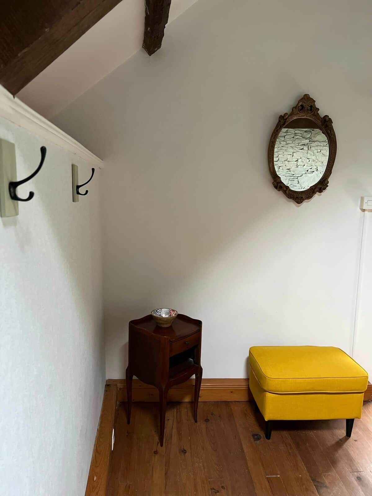Ein Zimmer mit Holzboden, einem kleinen Holztischchen mit einer Schale, einem gelben Hocker und einem an der Wand befestigten Spiegel.