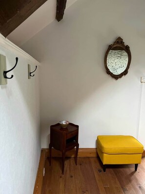 Een kamer met een houten vloer, een klein houten bijzettafeltje met een schaal, een geel poefje en een spiegel aan de muur.