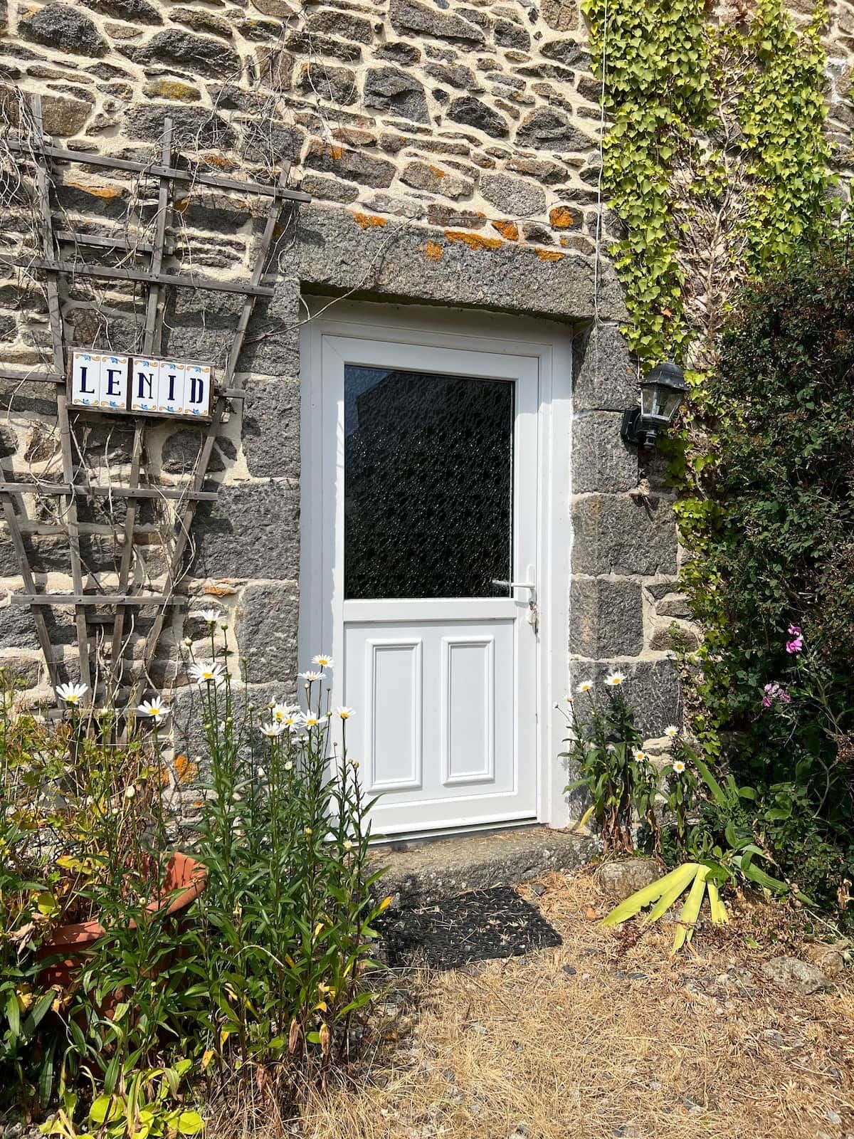 Un bâtiment en pierre avec une porte blanche, une enseigne en bois sur laquelle on peut lire « LENID », et un petit jardin agrémenté de fleurs et de plantes.