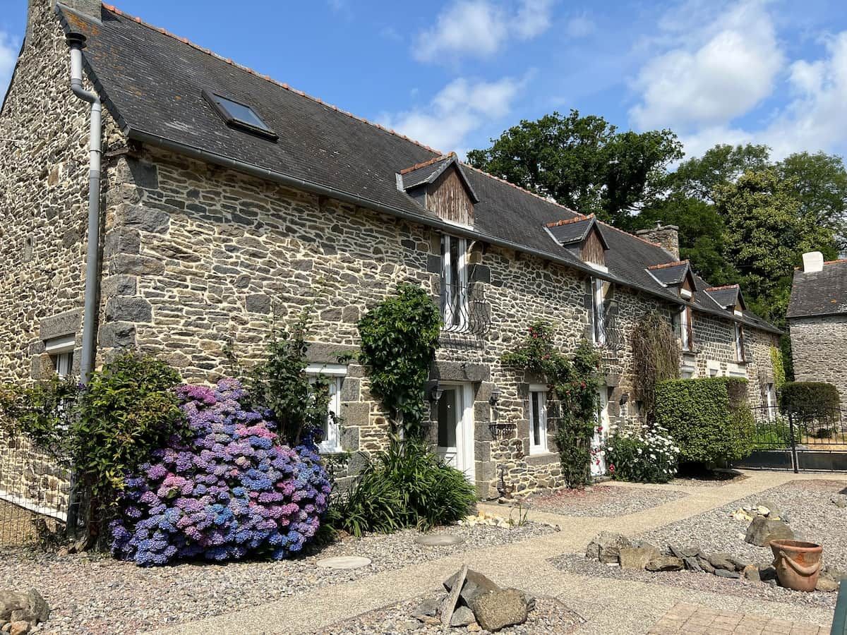 A stone building with a gravel driveway and a large bush of purple flowers.