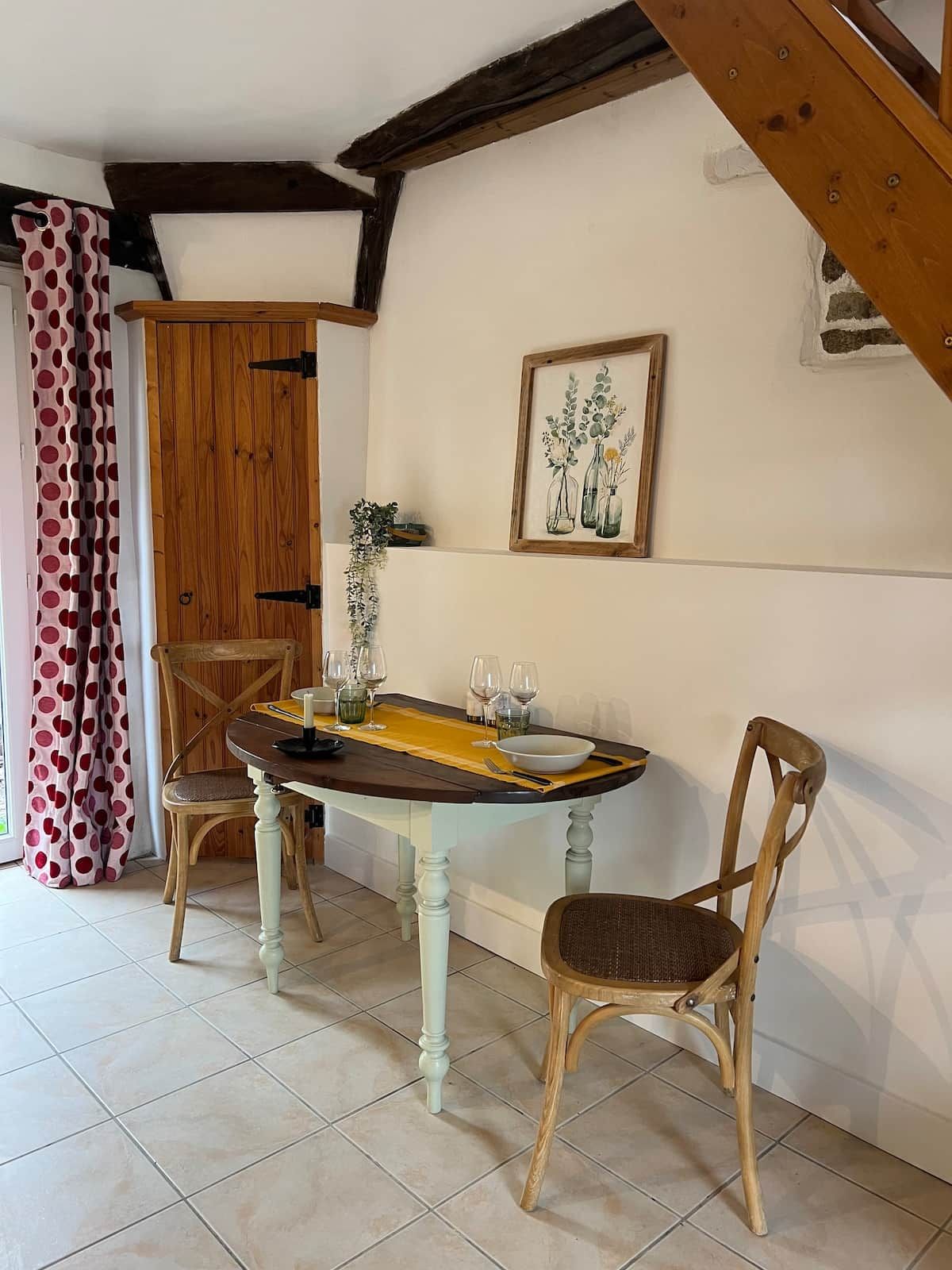 Un coin repas avec une table en bois, deux chaises, une estampe botanique encadrée et un rideau à motifs à pois.