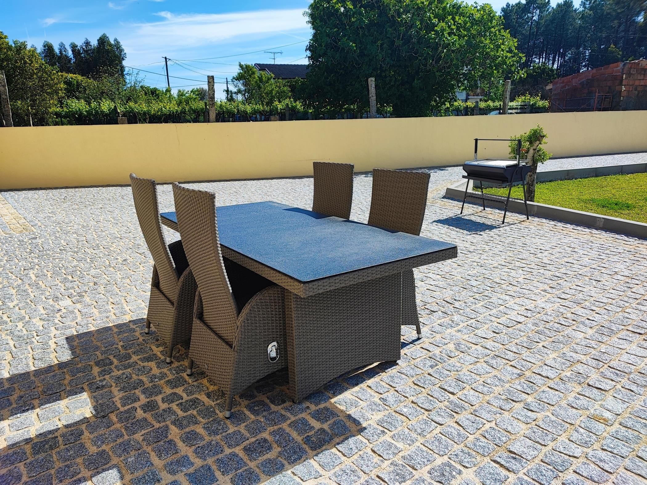 An outdoor patio with a table and chairs, a barbecue grill, and a paved area with trees and buildings in the background.