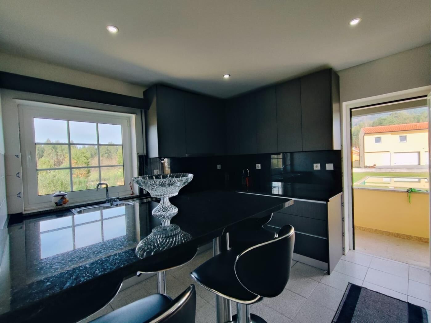 A modern kitchen with a black countertop, two bar stools, a crystal glass centerpiece, and a window with a view of greenery.