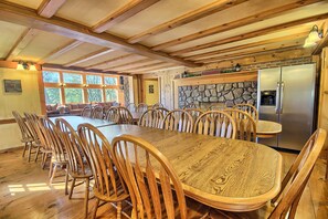 Una amplia mesa de comedor de madera rodeada de sillas en una habitación con chimenea de piedra y un refrigerador.