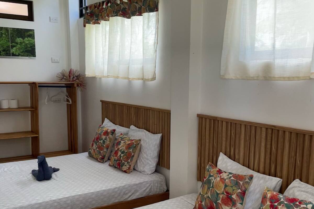 Une chambre d’hôtel avec deux lits, chacun doté d’une tête de lit en bois, de draps blancs et d’oreillers à motifs floraux. On y trouve également une étagère en bois avec des cintres et une petite plante.