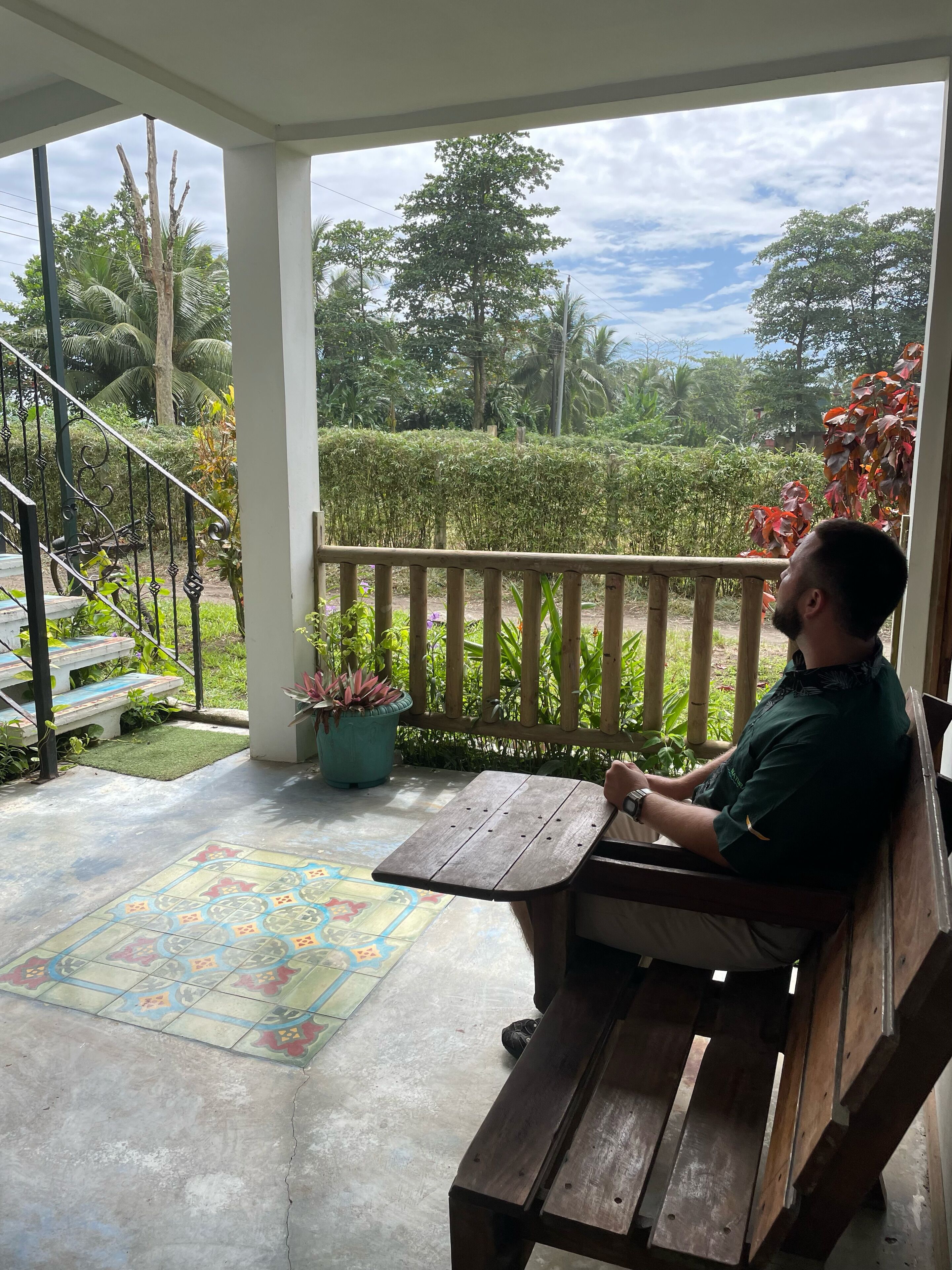 A man sitting on a wooden bench at a covered outdoor area with a view of a garden and trees.