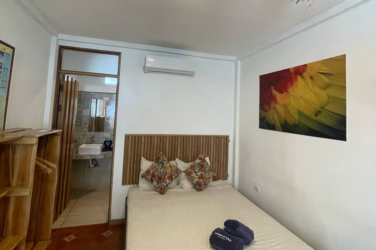 Une chambre d’hôtel avec un lit, un banc en bois, une salle de bain visible à travers une porte ouverte et un tableau accroché au mur.