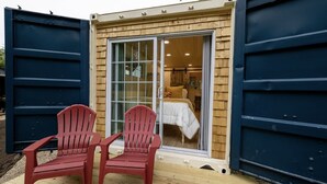 Una cabaña pequeña con una puerta corrediza de vidrio que da a un dormitorio con cama y mesita de noche. Hay dos sillas Adirondack rojas afuera de la cabaña.