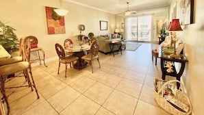 A spacious living area with a dining table, chairs, a sofa, and a coffee table. There is a large window providing natural light and a view of the outdoors. - #631 Beautiful poolside Condo perfect for a family getaway (Harrison County)
