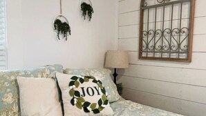A bedroom with a floral-patterned bedspread, a pillow with 'JOY' embroidered, a window with decorative iron bars, and two hanging plants.