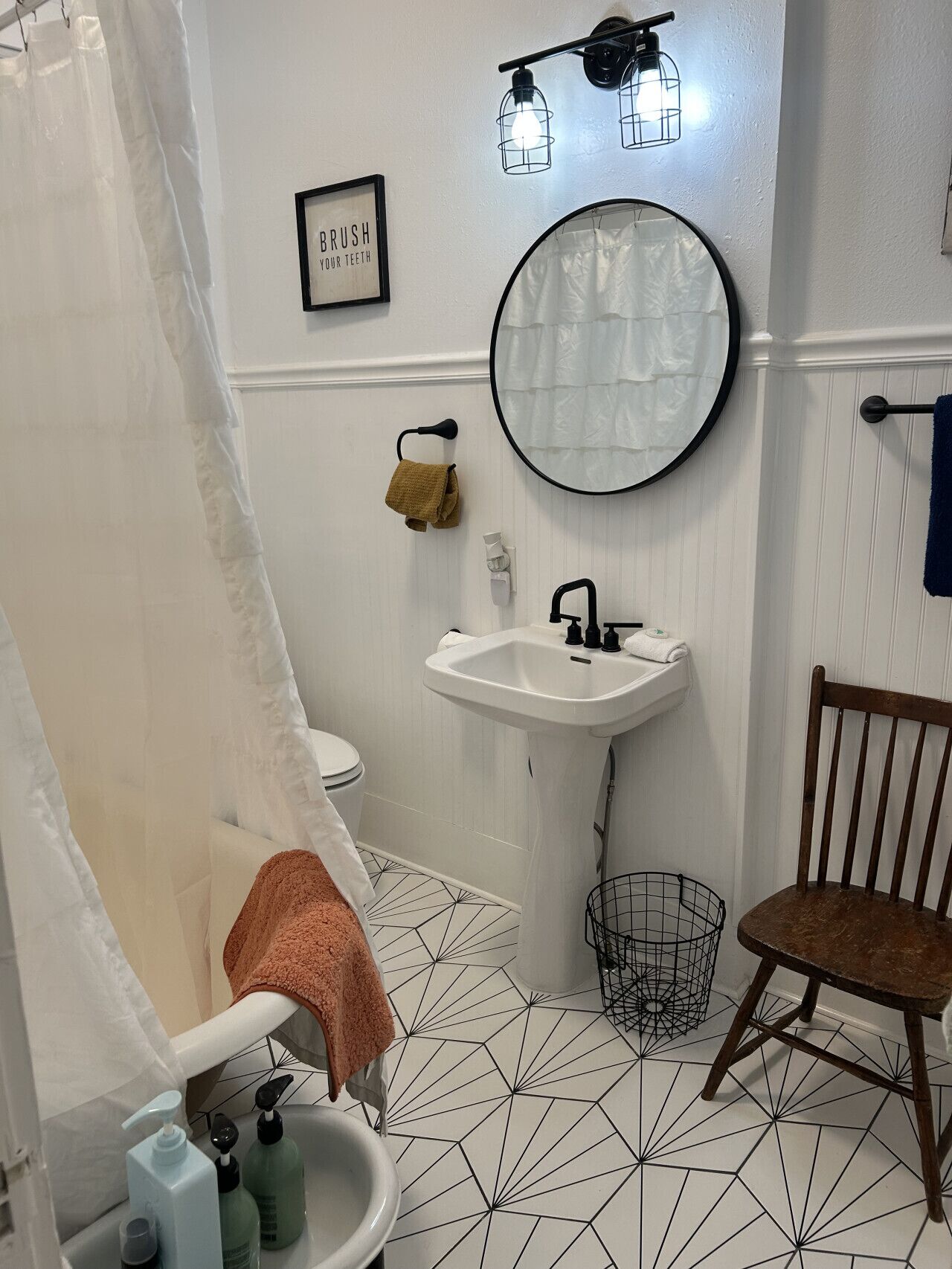 A bathroom with a white sink, a mirror, a towel rack, a wooden chair, and a geometric-patterned floor.