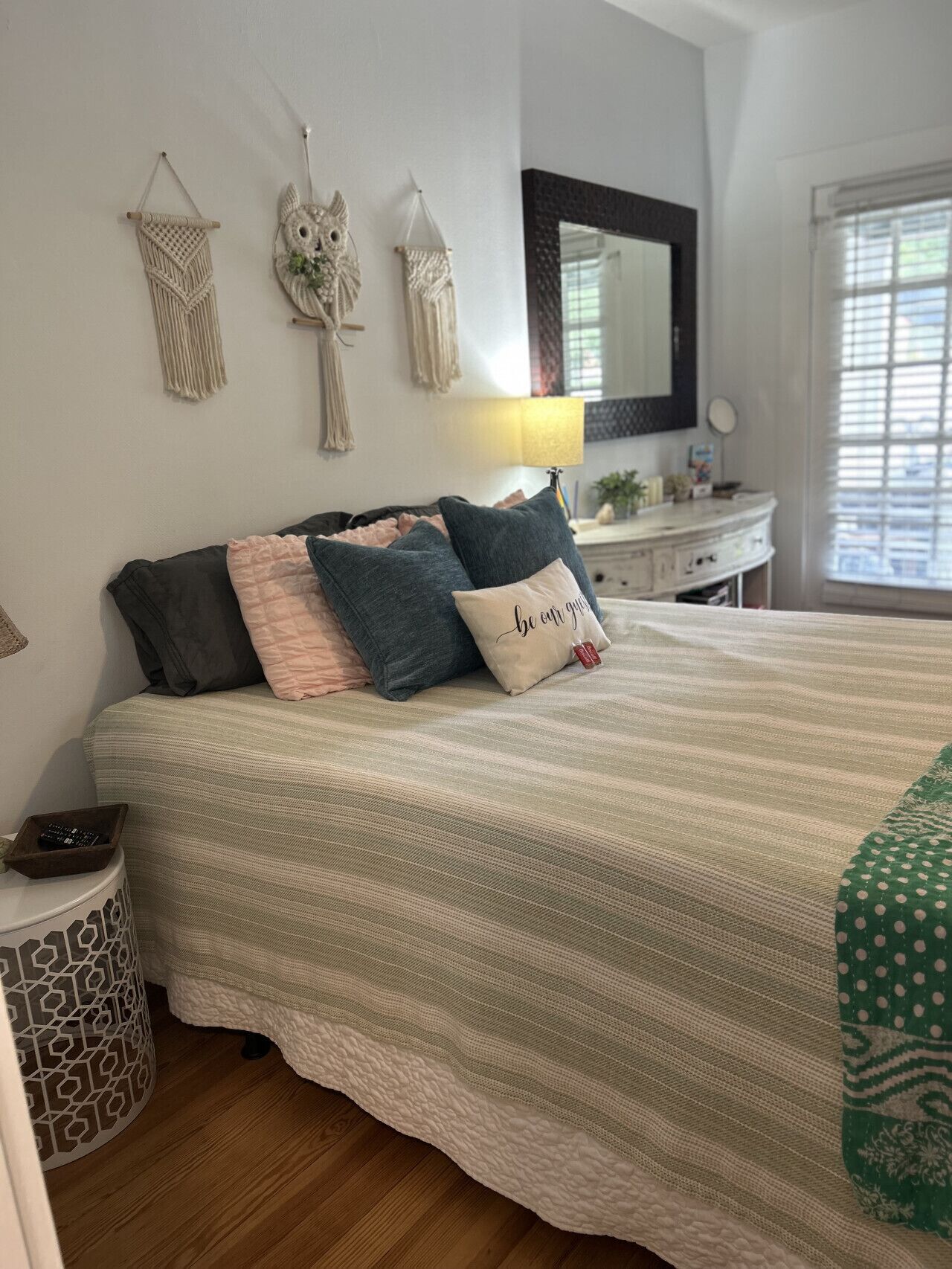 A neatly made bed with a striped bedspread, a bedside table with a lamp, and wall decor including macramé hangings.