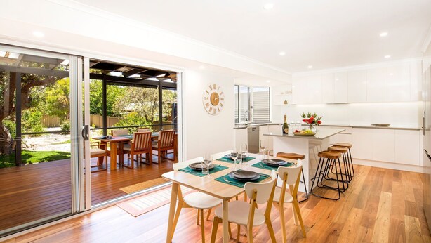 A modern kitchen with white cabinets, a dining area with wooden furniture, and a glass door leading to an outdoor patio. - Banksia by the Bay, Jervis Bay (Vincentia)