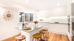 A modern kitchen with white cabinetry, a wooden island with stools, a large clock, and a window with a view. - Banksia by the Bay, Jervis Bay (Vincentia)