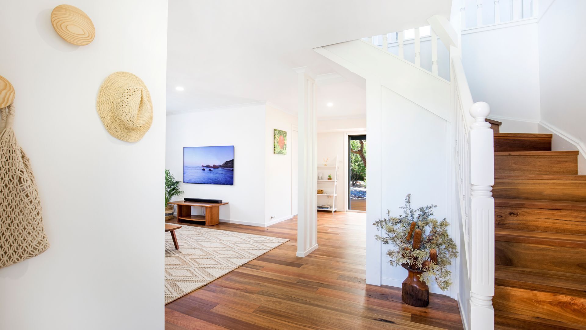 Un vaste salon doté d’un plancher en bois, d’un téléviseur fixé au mur, d’un escalier avec des rampes blanches et d’une vue sur l’extérieur grâce à une porte ouverte.