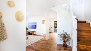 A spacious living room with a wooden floor, a TV mounted on the wall, a staircase with white railings, and a view of the outdoors through an open door. - Banksia by the Bay, Jervis Bay (Vincentia)