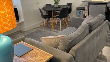 A modern living room with a grey sofa, a wooden coffee table, a dining table with chairs, and a kitchen area with cabinets and appliances.