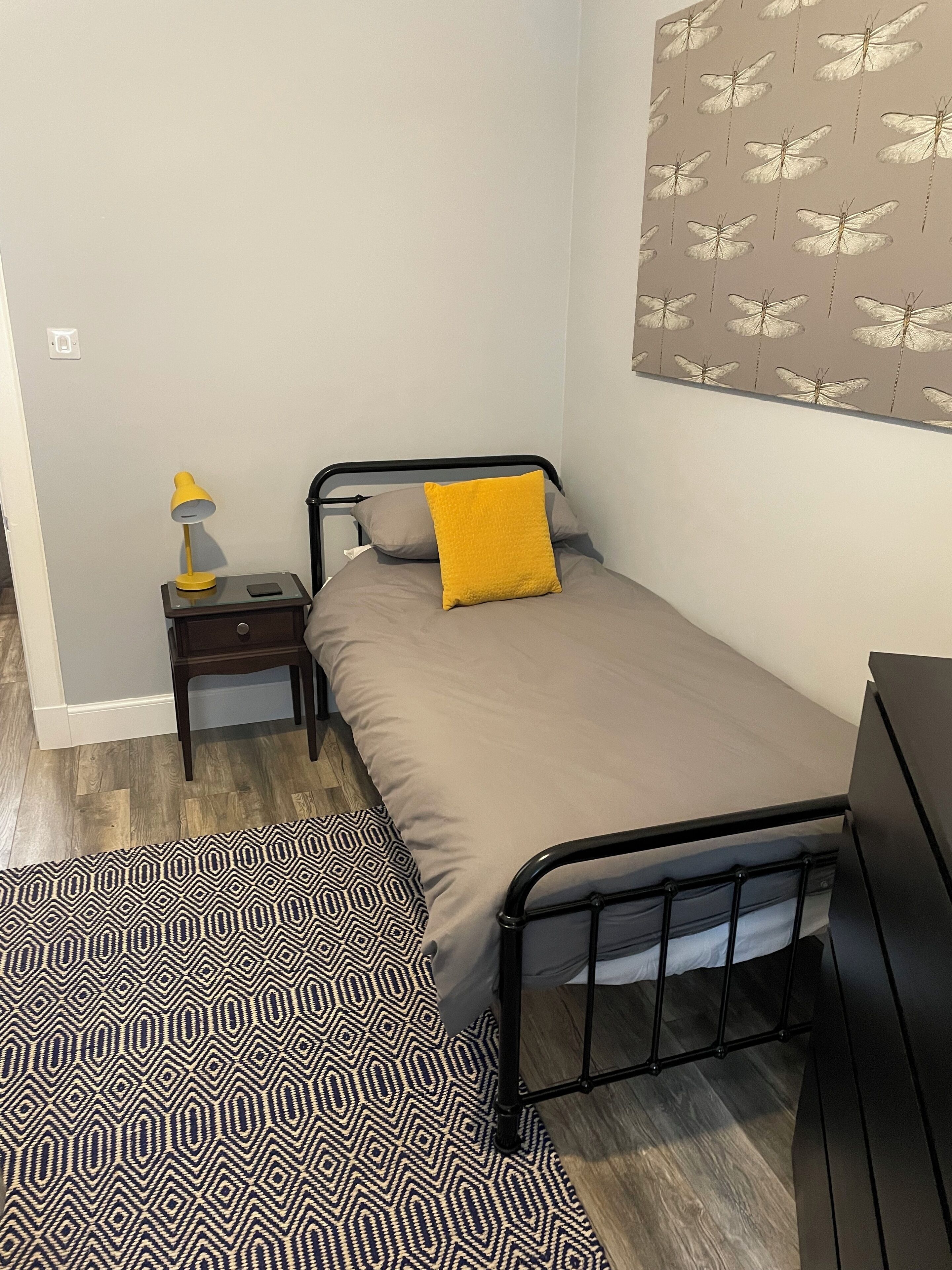 Ein kleines, ordentlich eingerichtetes Schlafzimmer mit Bett, Nachttisch, Lampe und einem gemusterten Teppich.