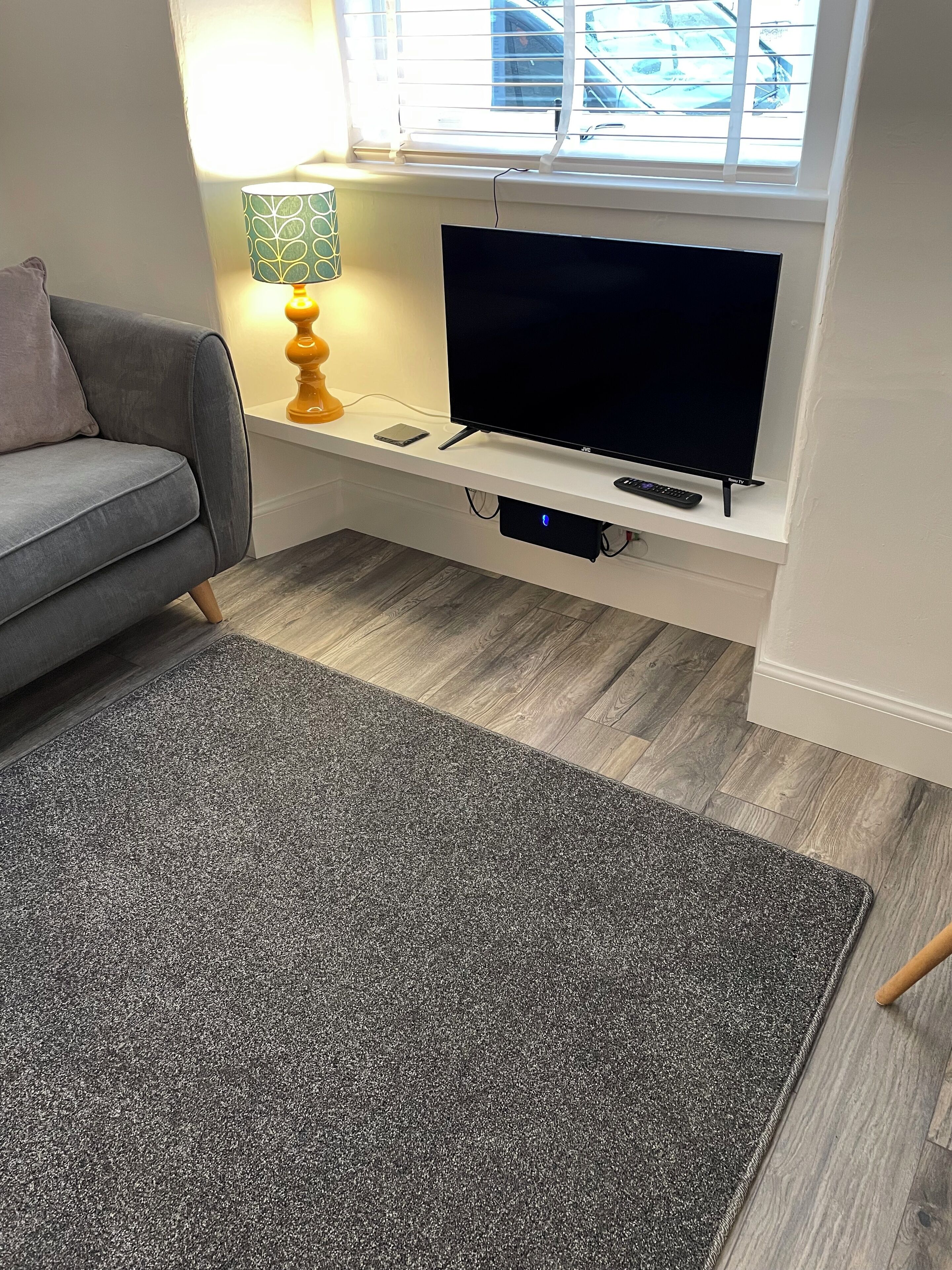 A living room with a grey sofa, a flat-screen TV on a white stand, a small table with a lamp, and a grey rug.