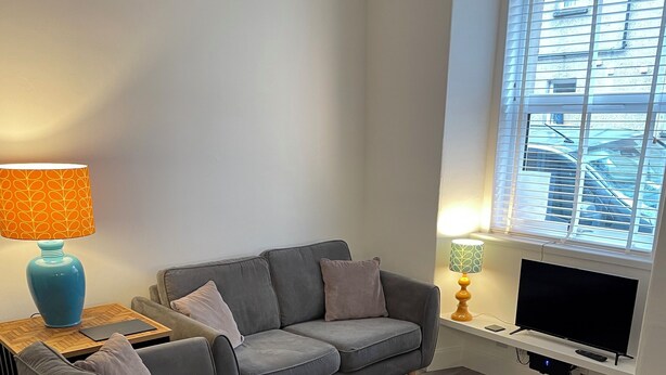 A modern living room with a grey sofa, a small wooden side table, a blue lamp, and a TV on a stand. - No.4 May Terrace (Inverness)