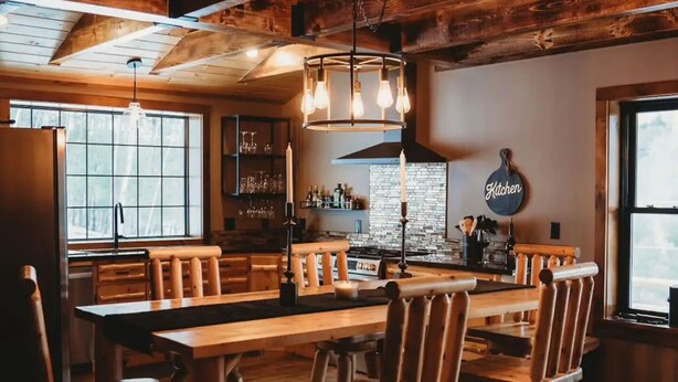 Un interior rústico de madera con una gran mesa de comedor y sillas, una zona de cocina con una lámpara colgante y un letrero que dice 'Kitchen'.