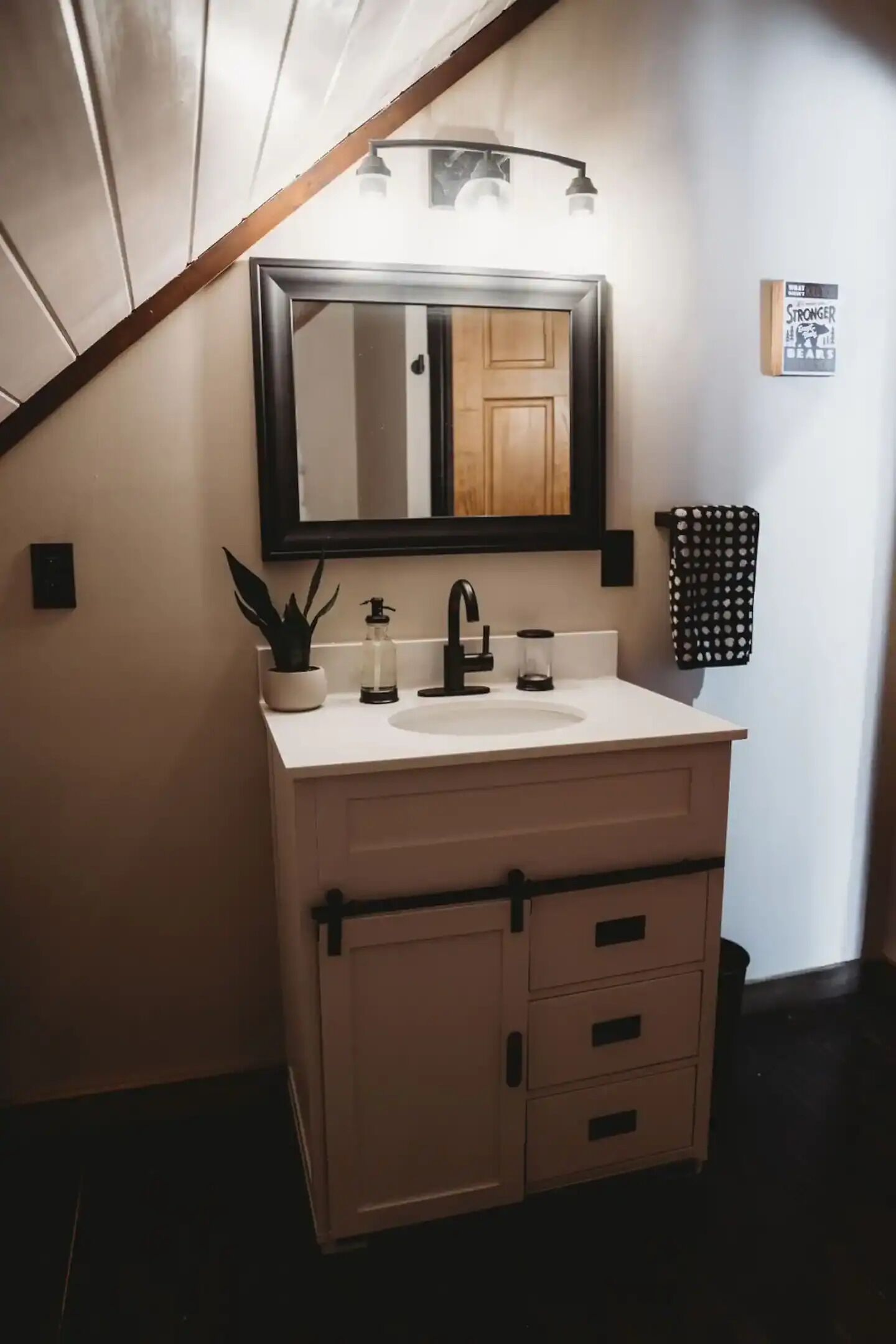 A bathroom with a white vanity, black faucet, and a mirror above the sink. There is a potted plant on the vanity and a towel rack with a polka dot towel.