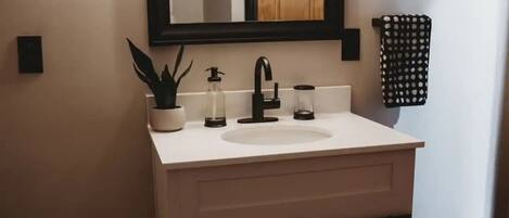 A bathroom with a white vanity, black faucet, and a mirror above the sink. There is a potted plant on the vanity and a towel rack with a polka dot towel.
