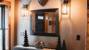 Techo de madera con vigas a la vista, un espejo fijado a la pared, dos apliques, una decoración con un pequeño árbol y un mueble con tela blanca.
