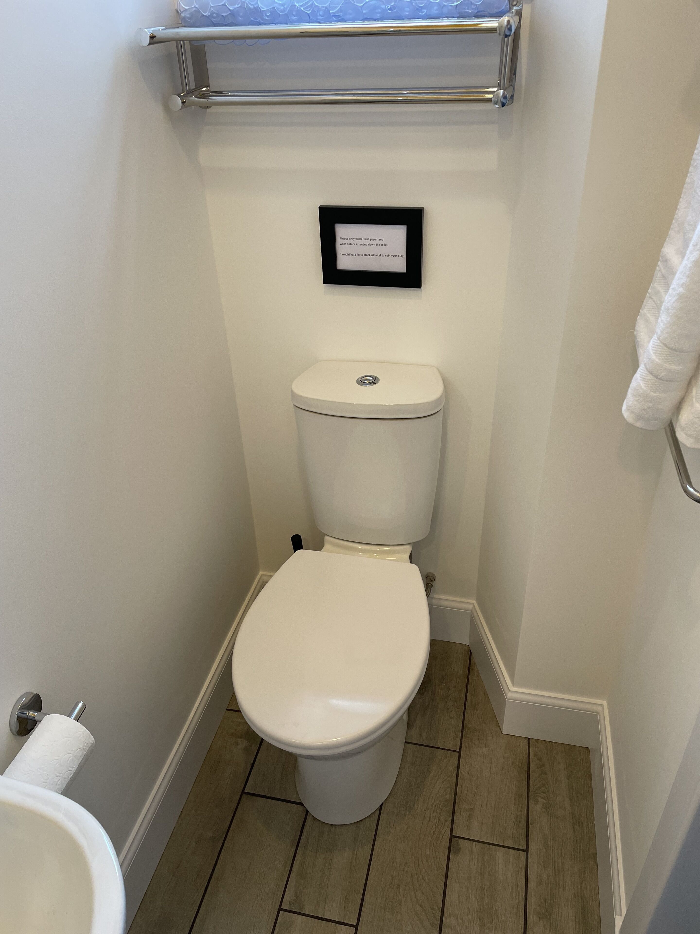 De petites toilettes blanches, dotées d’une armature en métal, d’un porte-rouleau de papier toilette blanc et d’une affiche encadrée accrochée au mur.