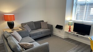 A modern living room with a grey sofa, a small wooden table, a TV on a stand, and a window with blinds. - City Apartment - No.1 May Court (Inverness)