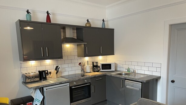 A compact kitchen with grey cabinets, a microwave, a toaster, and a coffee maker. There is a small trash bin and a grey sofa with a cushion. - City Apartment - No.1 May Court (Inverness)