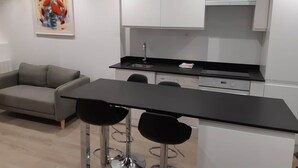Una cocina moderna con armarios blancos, una encimera negra y un sofá gris. Hay dos taburetes negros y un cuadro en la pared.