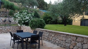 Un patio al aire libre con muro de piedra, una mesa con sillas y diversas plantas y árboles.