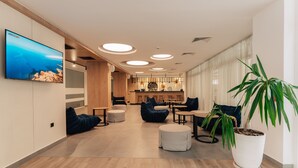 A modern interior with a wooden floor, a large TV on the wall, round ceiling lights, a central seating area with sofas and ottomans, and a potted plant. - TUI SUNEO PERLA ADULTS ONLY (Sunny Beach)