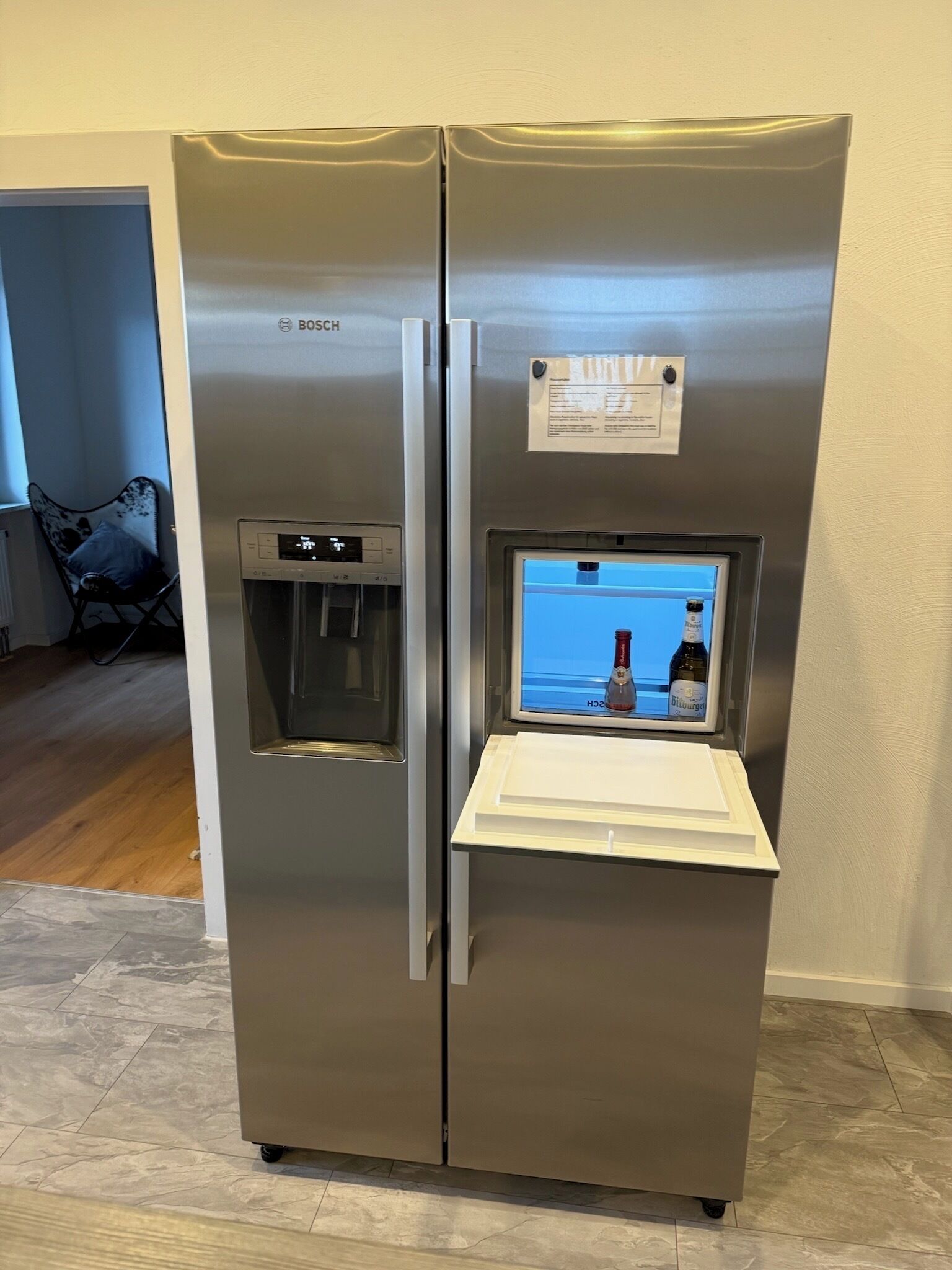 A stainless steel Bosch refrigerator with a built-in water dispenser and a small fridge compartment displaying beverages.