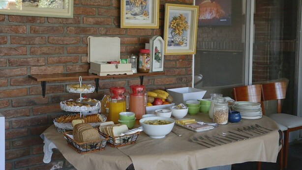 A breakfast buffet with various food items and drinks, set against a brick wall with framed artwork. - Posada Bernies (Ezeiza)