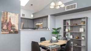 A modern kitchen with grey cabinets, a dining area with a glass table, black chairs, and a wooden shelf with decorative items. - Casas Adobes Summer Townhouse | Pool & Hot Tub (Tucson)