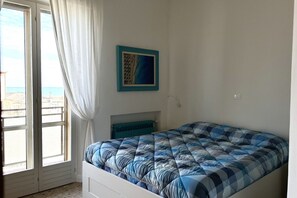 A bedroom with a bed, a window with curtains, a wall-mounted picture, and a view of the sea.