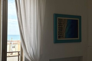 Una habitación con vista al mar a través de una ventana, un cuadro enmarcado en la pared y una cortina entreabierta.