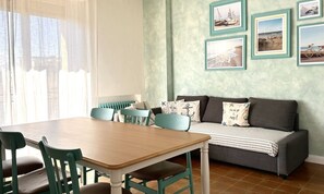 A dining area with a wooden table and chairs, a sofa with cushions, and a wall with framed pictures.