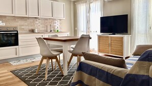 A modern kitchen with white cabinets, a dining table with chairs, a TV, and a sofa with a striped blanket. - P016 - Porto Recanati, new three-room penthouse 30m from the sea (Porto Recanati)
