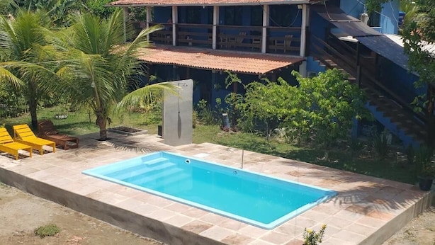 A swimming pool with a tiled deck, surrounded by greenery and a building with a red-tiled roof. - Pousada Tulum (Maraú)