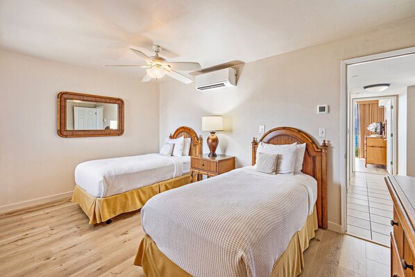 A hotel room with two beds, a ceiling fan, an air conditioning unit, a mirror, a nightstand with a lamp, and a wooden dresser.