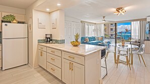 Una cocina moderna con gabinetes blancos, una isla central y una zona de comedor con sillas traslúcidas y una mesa de vidrio.