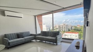 A modern living room with a sectional sofa, a small table, and a large window offering a city view. - Departamento a 20 metros de 5a Av. (Playa del Carmen)