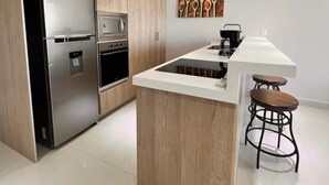 A modern kitchen with a stainless steel refrigerator, oven, and microwave. There is a wooden island with a sink and stools. - Departamento a 20 metros de 5a Av. (Playa del Carmen)