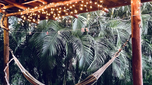Una hamaca debajo de una estructura de madera con luces colgantes, rodeada de exuberantes palmeras.