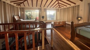 Una habitación amplia con vigas de madera en el techo, un ventanal con cortinas, una cama, un banco de madera y una mesa.