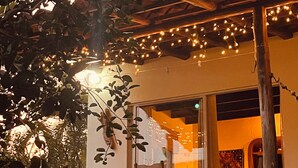 Un patio exterior con mobiliario de madera, luces colgantes y vista al interior a través de una puerta de cristal.